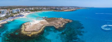 Turkuaz sular ve Nissi Sahili 'nin kumlu kıyıları, Ayia Napa, lüks tatil beldeleri, güneşlik yatakları, kayalık bir yarımada ve su aktivitelerinden hoşlanan insanlar..