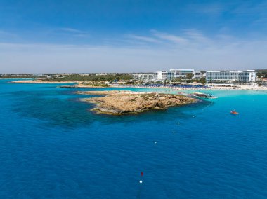 Kıbrıs Rum Kesimi 'nin Ayia Napa kentindeki Nissi Plajı' nın turkuaz sular, kumlu kıyılar, kayalık çıkıntılar, sahil otelleri ve deniz manzaralı havadan manzarası..
