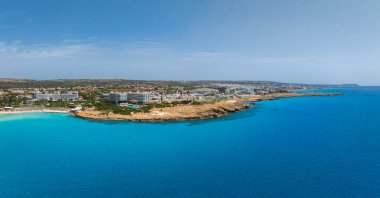 Kıbrıs Rum Kesimi 'nin Ayia Napa kentinin hava manzarasında turkuaz sular, Nissi Plajı, lüks tatil beldeleri, kumlu plajlar, kayalık çıkıntılar ve güneş ışığındaki kentsel alanlar görülüyor..
