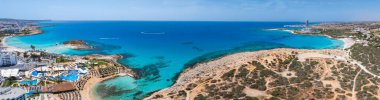 Kıbrıs Rum Kesimi 'nin Ayia Napa kentinin havadan görünüşü Nissi ve Landa sahillerini, turkuaz suları, lüks tatil beldeleri, yemyeşil otlaklar ve denizde bir tekne gösteriyor..