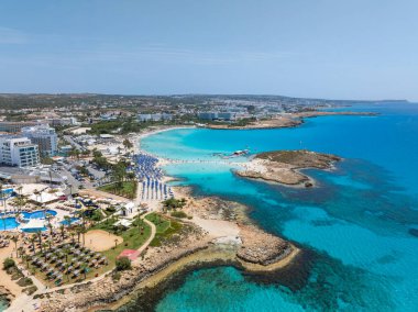 Kıbrıs 'ın Ayia Napa kentinin hava manzarasında turkuaz sular, Nissi ve Landa plajları, lüks tatil beldeleri, plaj şemsiyeleri ve yüzen insanlar yer alıyor..