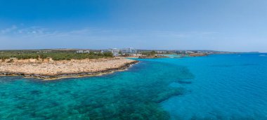 Kıbrıs Rum Kesimi 'nin Ayia Napa kentinin hava manzarası turkuaz sular, kayalık kıyı şeridi, kumlu plajlar, lüks oteller, yemyeşil alanlar ve açık mavi bir gökyüzünü gösteriyor..