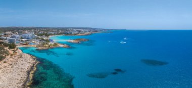 Kıbrıs Rum Kesimi 'nin Ayia Napa kentinin havadan görünüşü, Nissi Sahili' ni, turkuaz suları, lüks tatil beldeleri, şemsiyeli kumlu kıyıları ve sakin denizde kayıkları gösteriyor..
