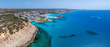 Kıbrıs Rum Kesimi 'nin Ayia Napa kentinin hava manzarasında turkuaz sular, kumlu plajlar, lüks tatil beldeleri, güneş pansiyonları, yemyeşil alanlar ve denizde bir sürat teknesi görülüyor..