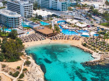 Kıbrıs 'ın Ayia Napa kentindeki turkuaz körfezi, kumlu plajlar, kayalık çıkıntılar, lüks tatil beldeleri, yüzme havuzları, palmiye ağaçları ve temiz sularda yüzen insanlarla dolu..