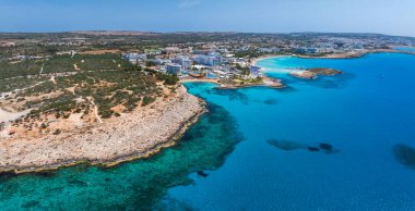 Kıbrıs Rum Kesimi 'nin Ayia Napa kentinin turkuaz suları, kumlu Nissi Sahili, lüks tatil beldeleri, engebeli kıyı şeridi ve yeşillikli kentsel yerleşim yerlerini gösteren hava görüntüleri..