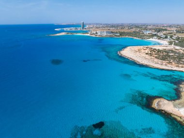 Kıbrıs Rum Kesimi 'nin Ayia Napa kentinin hava manzarasında turkuaz sular, kumlu plajlar, lüks tatil beldeleri, yüksek modern bir bina ve doğal bitki örtüsü görülüyor..