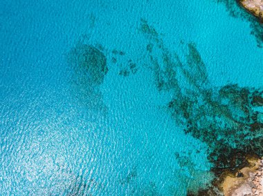 Kıbrıs Rum Kesimi 'nin Ayia Napa kenti yakınlarındaki turkuaz sularda, sualtı özelliklerini gösteren koyu renkli yamalar görülüyor. Sağ üst köşede engebeli, kayalık bir kıyı şeridi görünüyor..