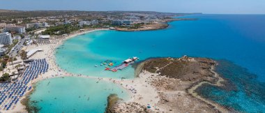 Kıbrıs Rum Kesimi 'nin Ayia Napa kentindeki Nissi Sahili' nin hava manzarasında turkuaz sular, kumlu kıyılar, güneş pansiyonları, yüzücüler, tatil beldeleri ve yemyeşil alanlar görülüyor..