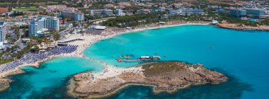 Kıbrıs Rum Kesimi 'nin Ayia Napa kentindeki Nissi Plajı' ndaki turkuaz sular, kumlu kıyılar ve lüks tatil beldeleri. Yüzücüler, su araçları ve kayalık bir ada canlı sahneyi canlandırıyor..