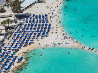 Kıbrıs Rum Kesimi 'nin Ayia Napa kentindeki Nissi Plajı' nın hava manzarasında turkuaz sular, kumlu kıyılar, mavi şemsiyeler, güneş panzeleri ve yakınlardaki tatil köyleri görülüyor..