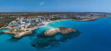 Ayia Napa, Kıbrıs Rum Kesimi, Nissi Sahili, turkuaz sular, altın kumlar, lüks tatil beldeleri, yemyeşil alanlar ve yüzen insanların yer aldığı hava manzarası.