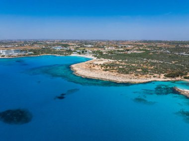 Kıbrıs Rum Kesimi 'nin Ayia Napa kentinin hava manzarasında turkuaz sular, kayalık kıyılar, dağınık binalar, tatil beldeleri, yemyeşil alanlar ve açık mavi bir gökyüzü görülüyor..