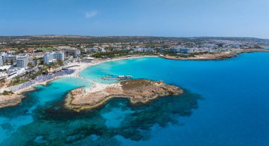 Kıbrıs Rum Kesimi 'nin Ayia Napa kentindeki Nissi Plajı' nın hava manzarasında turkuaz sular, kumlu kıyılar, lüks tatil beldeleri, güneş pansiyonları ve yüzen insanlar yer alıyor..