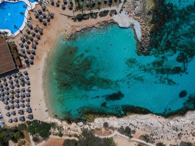 Kıbrıs 'taki Ayia Napa plajının hava görüntüleri, turkuaz sular, kumlu kıyılar, güneş pansiyonları, şemsiyeler, tatil beldesi ve görünür bir yüzme havuzu yer alıyor..