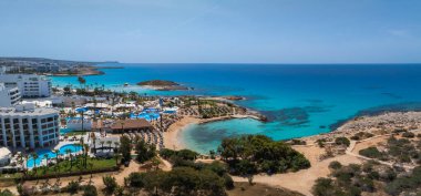 Kıbrıs Rum Kesimi 'nin Ayia Napa kentinin havadan görünüşü, Nissi Sahili' ni, turkuaz suları, lüks tatil beldeleri, yüzme havuzları, güneşlik salonları, kayalık bir ada ve yemyeşil yemyeşil alanları gösteriyor..
