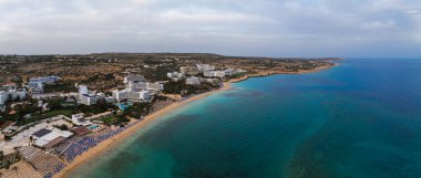 Kıbrıs Rum Kesimi 'nin Ayia Napa kentinin hava manzarasında turkuaz sular, kumlu plajlar, lüks tatil beldeleri, plaj şemsiyeleri ve kentsel ve doğal manzaraların karışımı görülüyor..