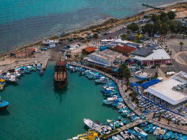 Aerial image of Ayia Napa marina in Cyprus featuring a wooden ship, small boats, turquoise Mediterranean waters, rocky shore, and a circular fountain.