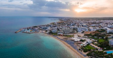 Kıbrıs Rum Kesimi 'nin Ayia Napa kentinin hava manzarasında turkuaz sular, mavi şemsiyeli kumlu plajlar, gün batımında tekneli bir marina ve kentsel alanlar görülüyor..