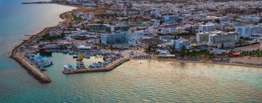 Kıbrıs Rum Kesimi 'nin Ayia Napa kentinin hava görüntüsü, renkli tekneleri, mavi şemsiyeli kumlu plajları, turkuaz suları ve beyazlatılmış kentsel binaları gösteriyor..