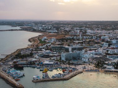 Kıbrıs Rum Kesimi 'nin Ayia Napa kentinin hava manzarasında, gün batımında tekneleri, kumlu plajları, berrak suları, lüks otelleri ve kentsel yeşillikleri olan canlı bir marina görülüyor..