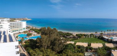 Kıbrıs Rum Kesimi 'nin Ayia Napa kentinin hava manzarası, lüks tatil beldeleri, kumlu plajlar, temiz mavi sular, güneş pansiyonları, şemsiyeler ve eğimli bir kıyı şeridini gösteriyor..