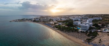 Kıbrıs Rum Kesimi 'nin Ayia Napa kentinin hava manzarası, Nissi Sahili' ni, lüks otelleri, turkuaz Akdeniz sularını ve gün batımında kentsel manzarayı gösteriyor.
