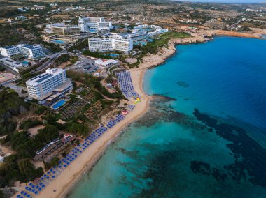 Kıbrıs Rum Kesimi 'nin Ayia Napa kentinin havadan görünüşü, turkuaz sular, kumlu plajlar, lüks tatil beldeleri, mavi şemsiyeler, yemyeşil ve kentsel kırsal manzaraları gösteriyor.