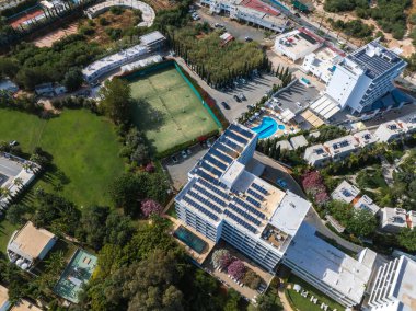 Kıbrıs, Ayia Napa 'nın hava görüntüsü, bir tenis kortu, güneş panelleri, yüzme havuzu, peyzaj bahçeleri ve yeşillik içeren modern beyaz bir bina gösteriyor..