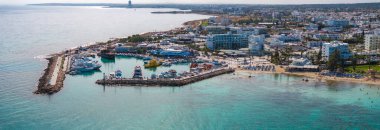Aerial view of Ayia Napa, Cyprus, showing a marina with boats, sandy beaches with sun loungers, turquoise waters, luxury hotels, and urban scenery.