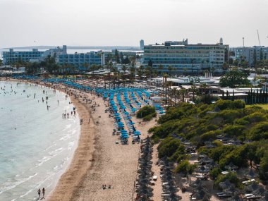 Kıbrıs Rum Kesimi 'nin Ayia Napa kentindeki kumlu bir sahil manzarası, mavi güneş pansiyonları, berrak sularda yüzücüler, lüks oteller ve arka planda yemyeşil alanlar..
