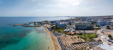 Kıbrıs Rum Kesimi 'nin Ayia Napa kentsel manzarası, turkuaz suları, güneş pansiyonlu kumlu plajlar, lüks oteller, marina ve güneşli gökyüzünün altındaki kentsel yerleşim yerlerini gösteriyor..