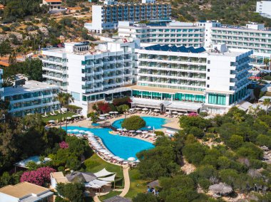 Balkonları geniş bir havuz, güneş pansiyonları ve yeşillik çevresindeki modern beyaz tatil köyleri binaları. Tepe manzarası ve bahçeler arka planda görünür..