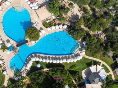 Kıbrıs Rum Kesimi 'nin Ayia Napa kentindeki bir tatil köyünün havuz görüntüsünde, ahşap bir köprü, beyaz şemsiyeler, güneş panzeleri, yemyeşil alanlar ve manzaralı bahçeler yer alıyor..