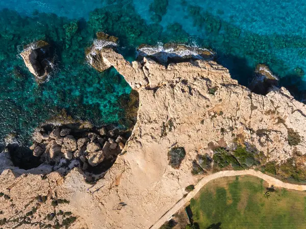 Kıbrıs Rum Kesimi 'nin Ayia Napa kentinin bej kaya oluşumları, turkuaz Akdeniz suları, yeşil bir çimen ve eğimli bir patika gösteren hava görüntüsü..