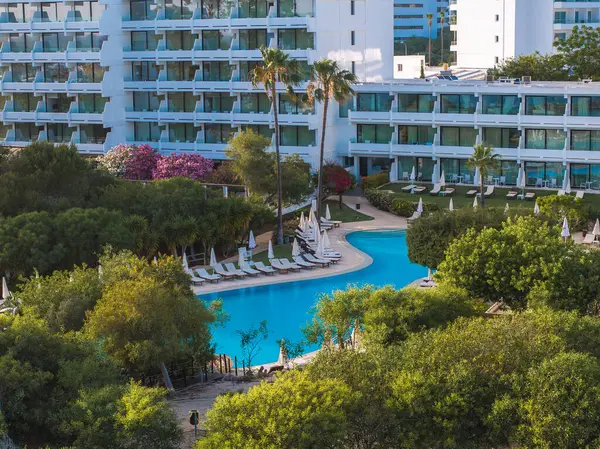 Kıbrıs Rum Kesimi 'nin Ayia Napa kentinde büyük bir havuz, şezlonglar, şemsiyeler, palmiye ağaçları, yemyeşil alanlar ve arka planda modern çok katlı bir otelin yer aldığı bir tatil beldesi..