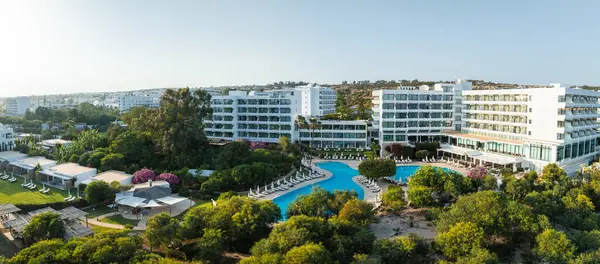 Modern lüks otel, yemyeşil, canlı çiçekler, ve dinlenme yerlerinin olduğu büyük bir havuz. Arka planda kentsel ve doğal manzaralar.