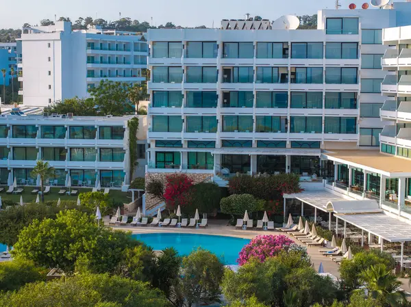 Camdan pencereleri ve balkonları olan modern çok katlı bir otel, güneşli havuzlu merkezi bir havuz, canlı bir yeşillik ve Ayia Napa 'da açık mavi bir gökyüzü..