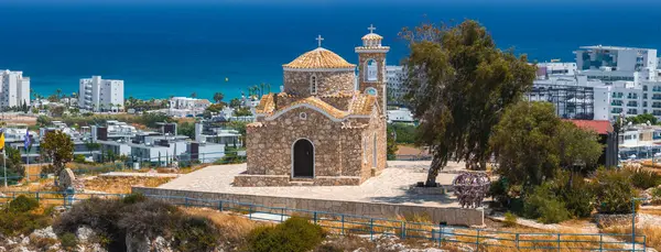 Taştan bir St. Elias kilisesi kiremit çatılı ve çan kulesiyle Kıbrıs 'ın Ayia Napa kentindeki bir tepede, modern binalar ve arka planda Akdeniz bulunur..