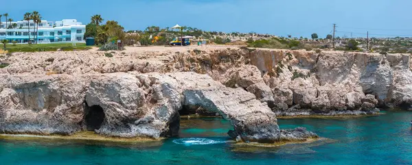 Kıbrıs Rum Kesimi 'nin Ayia Napa kentindeki turkuaz suların üzerinde doğal bir kaya sevgisi kemeri olan kireçtaşı kayalıkları. Yakınlarda bir tatil köyü, palmiye ağaçları ve park edilmiş bir araç görülüyor..