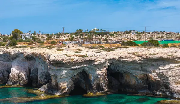 Deniz mağaralı kireçtaşı kayalıkları Kıbrıs Rum Kesimi 'nin Ayia Napa kentinde turkuaz sularla buluşuyor. Beyaz kubbeli bir yapı ve şehir manzarası güneşli gökyüzünün altında görülebilir..