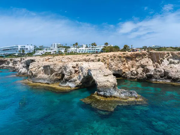 Kıbrıs Rum Kesimi 'nin Ayia Napa kentindeki turkuaz sulara uzanan doğal bir kaya kemeri, modern beyaz tatil beldeleri, palmiye ağaçları ve arka planda parlak mavi bir gökyüzüne sahiptir..