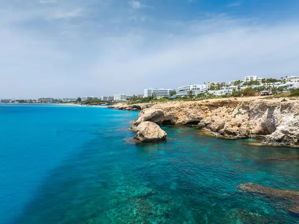 Kıbrıs Rum Kesimi 'nin Ayia Napa kentindeki turkuaz sular ve kayalık uçurumlar, modern beyaz lüks oteller ve arka planda açık mavi bir gökyüzünün altında yeşillik.