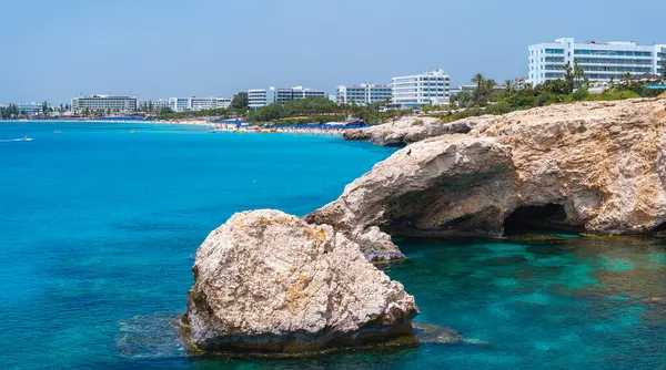 Kıbrıs 'taki Ayia Napa kıyı şeridi berrak turkuaz sular, engebeli kayalar, kumlu plajlar, güneşli banyolar ve arka planda modern lüks oteller.