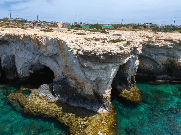 Doğal deniz mağaraları olan kayalık kayalıklar Kıbrıs Rum Kesimi 'nin Ayia Napa kentinde turkuaz sularla karşılaşıyor. Arka planda bir heykel parkı ve dağınık binalar görülüyor..