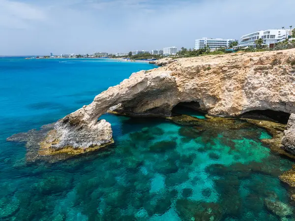 Kıbrıs Rum Kesimi 'nin Ayia Napa kentindeki turkuaz suların üzerinde doğal kaya aşk kemeri, görünür deniz yatakları ve arka planda kıyı şeridini kaplayan modern beyaz oteller bulunuyor..
