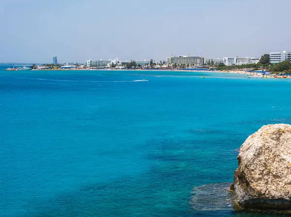 Kıbrıs Rum Kesimi 'nin Ayia Napa kentinin hava manzarasında turkuaz sular, Nissi Sahili, lüks tatil beldeleri, bir kaya oluşumu, küçük kayıklar ve açık gökyüzü altında yüzücüler görülüyor..