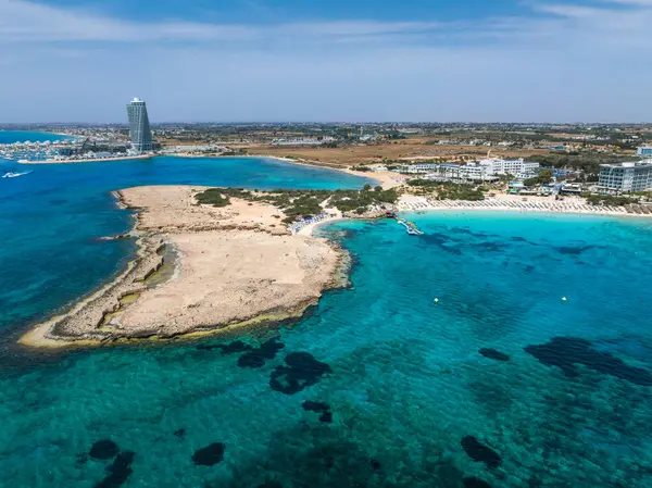 Kıbrıs Rum Kesimi 'nin Ayia Napa kentindeki Nissi Plajı' nın turkuaz sular, kumlu kıyılar, güneşli yataklar, şemsiyeler, lüks tatil beldeleri ve modern bir gökdelen binası ile hava manzarası.