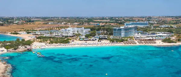Kıbrıs 'ın Ayia Napa kentinin hava manzarasında turkuaz sular, kumlu plajlar, lüks oteller, güneşlikler, şemsiyeler, küçük tekneler ve yüzen insanlar görülüyor..