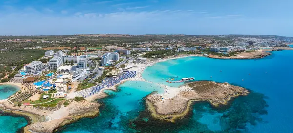 Kıbrıs Rum Kesimi 'nin Ayia Napa kentinin havadan görünüşü, Nissi Sahili, turkuaz sular, kumlu kıyılar, lüks tatil beldeleri, plaj şemsiyeleri ve denizde yüzücüler yer alıyor..